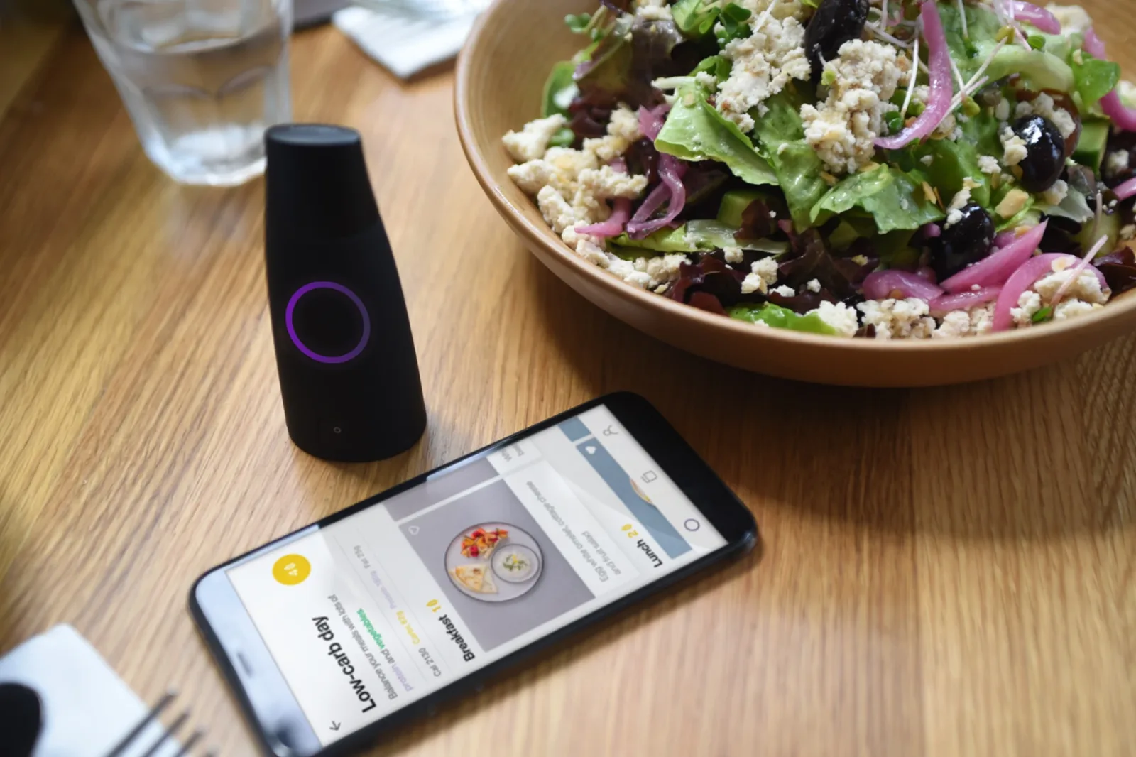 The Lumen metabolic breath device beside a phone showing a low-carb meal plan and a fresh salad